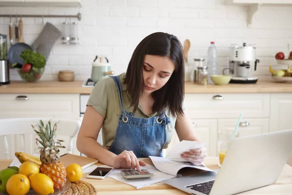 Portrait of young dark haired female wearing casual clothes at home, having focused concentrated look while calculating her debts for rent in kitchen, making notes, surrounded with papers and gadgets