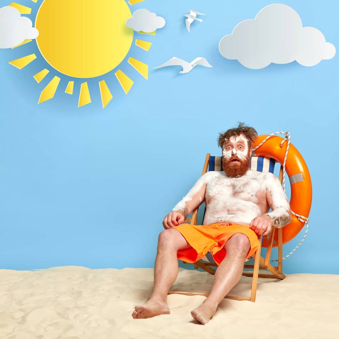 Shocked man has sunscreen cream on body to protect skin from uv rays, has summer vacation, burnt skin during sunbathinng, sits on comfortable beach chair, enjoys good rest during hot sunny day.